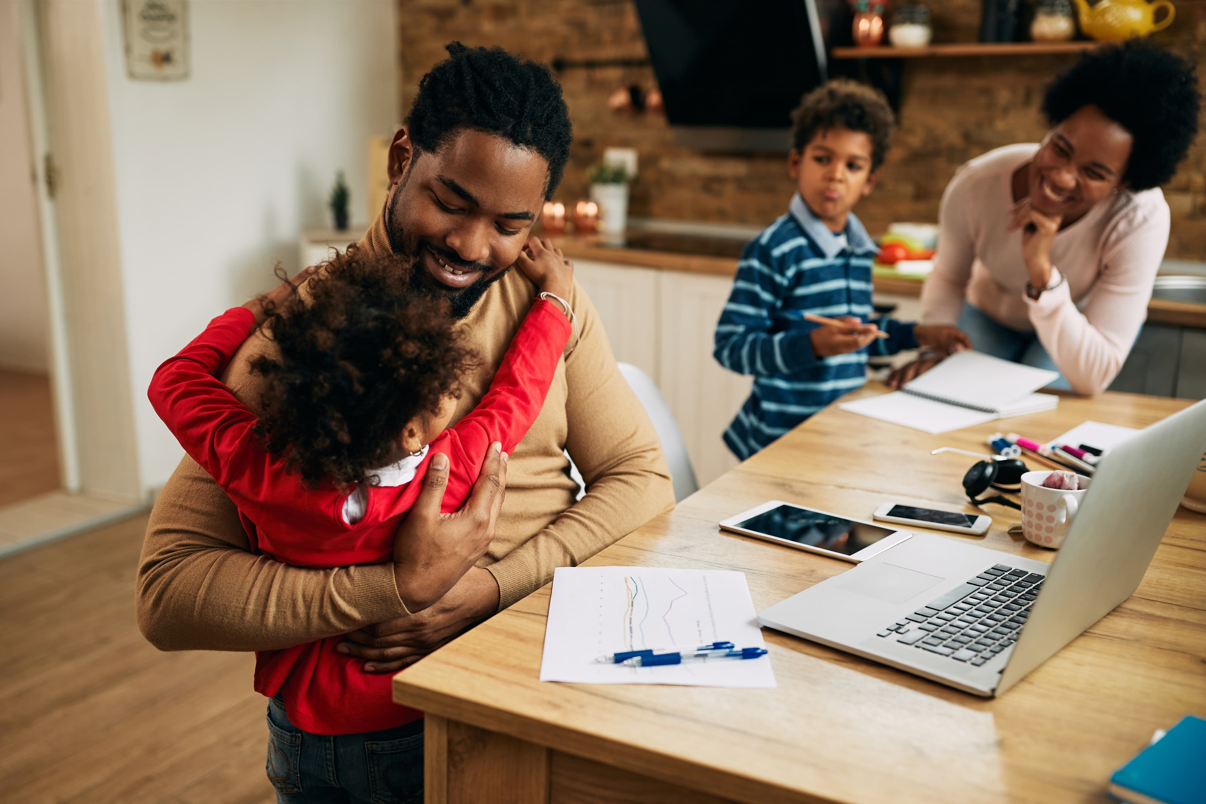 A father embracing his child at the end of a work day, with the rest of the family close by
